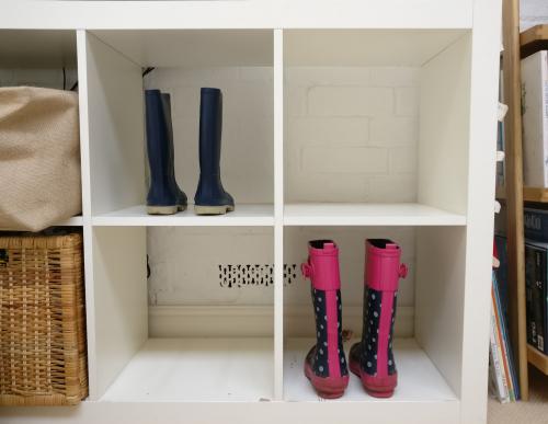 A white cube storage unit with a pair of pink wellies in one compartment and a pair of black wellies in another.