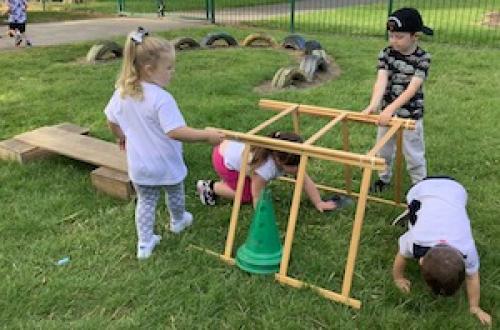Some children are crawling through a tunnel made of sticks. There are plastic cones and a wooden plank beside the tunnel.