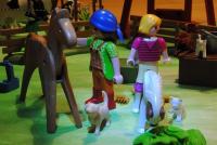 A green mat with some plastic toys placed on top of it: people, horses, cats and dogs. The toys are interacting with each other.