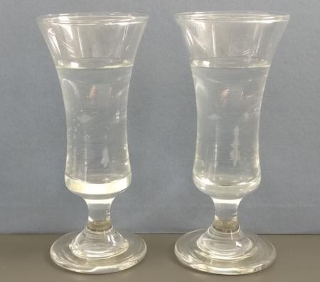Two glasses of water. The glasses curve outwards at the top. The water comes around three quarters of the way up in each glass.