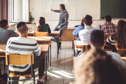 A classroom of older students at desks. There is a teacher sitting on a desk at the front of the room, pointing at the whiteboard behind him.