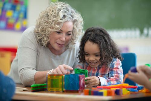 A teacher and a smiling young girl sitting at a table. The girl is manipulating construction materials.