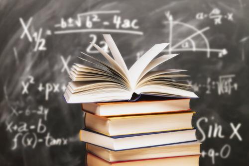 A stack of books, with the top one open, fanning pages. There is a blackboard in the background showing algebraic expressions and graphs.
