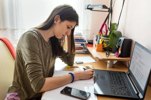 A young woman sitting at a desk writing on paper, with a laptop in front of her.