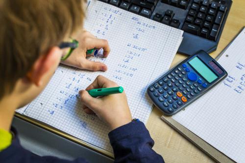 A boy writing with a pen on paper, with a calculator on his desk.