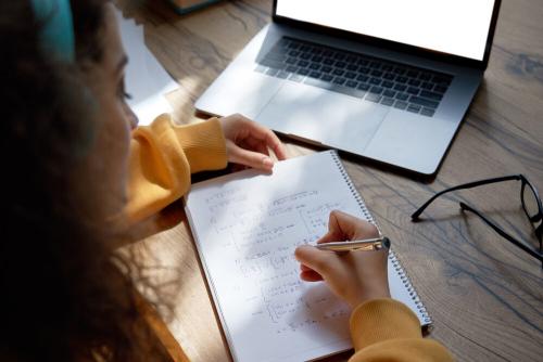 A woman sitting at a wooden desk. She is writing on a notepad with a laptop in the background.