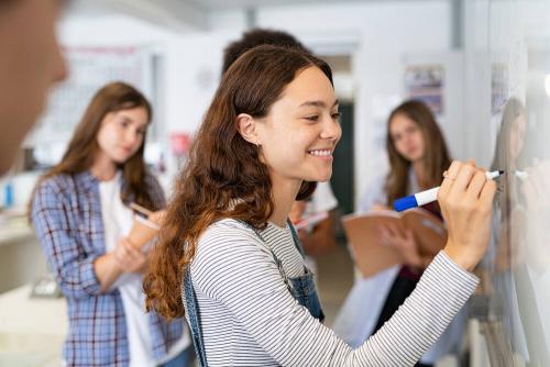 Several teenage students standing in front of a large whiteboard. One girl is writing on it and smiling.