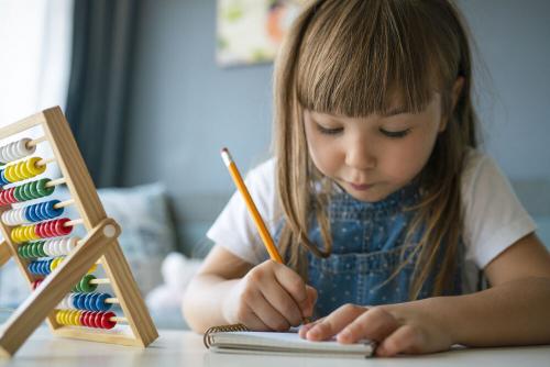 A young girl writing with a pencil. An abacus is on the table in front of her.