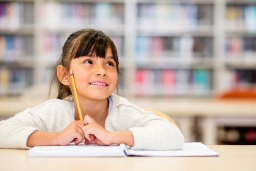Young girl smiling while looking upwards, holding a pencil and sitting at a table with a notebook.