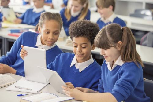 Three students sitting at a table, all looking at the same tablet and smiling.