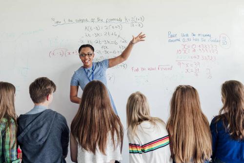 A teacher standing in front of a group of secondary students. The whiteboard behind them shows algebraic equations in a variety of colours.