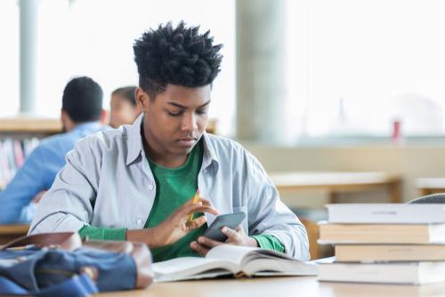 A teenage boy using his phone, sitting at a table covered with books.