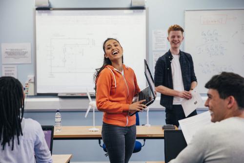 A teenage boy and girl standing at the front of a class. The girl is holding an open laptop and laughing.