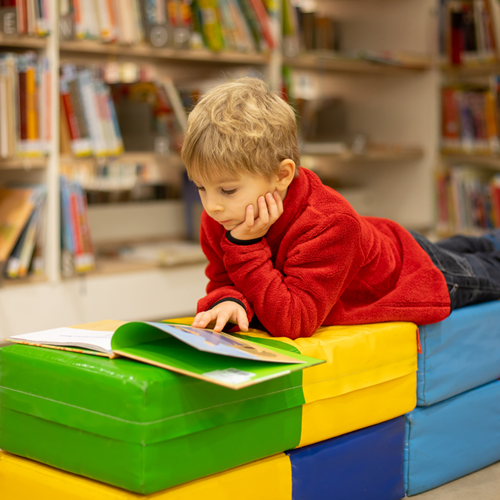 A boy lying on colourful blocks in a library, reading a book.