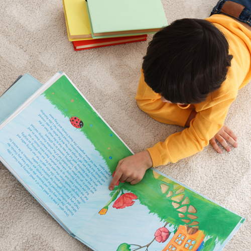 A little boy lying on his front, reading a book and pointing at the pictures.